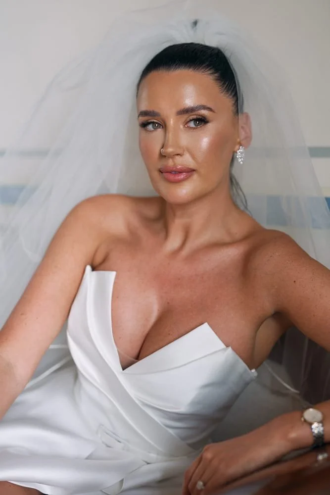 A woman dressed in a wedding gown with a sheer veil, wearing earrings and a ring, sitting and taking a selfie.