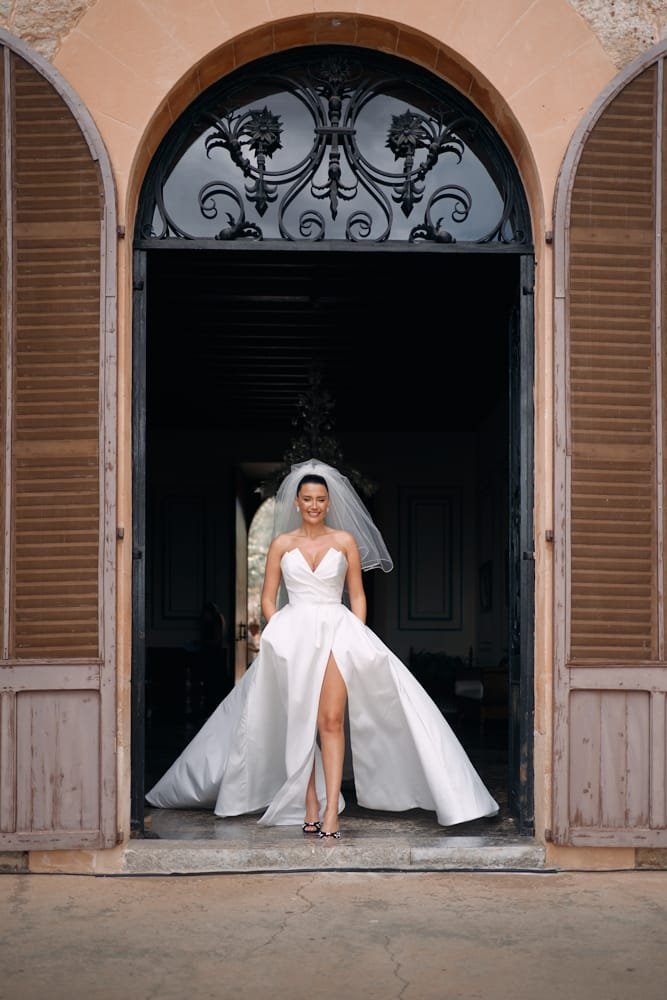 A bride in a white wedding dress with a thigh slit, standing in the doorway of a building with wooden shutters and decorative ironwork above the door, smiling and showing her high heel shoes.