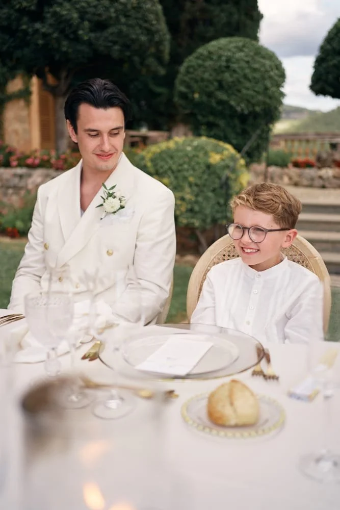 A man and a young boy smiling at each other at an outdoor table during a celebration, with a green garden in the background.