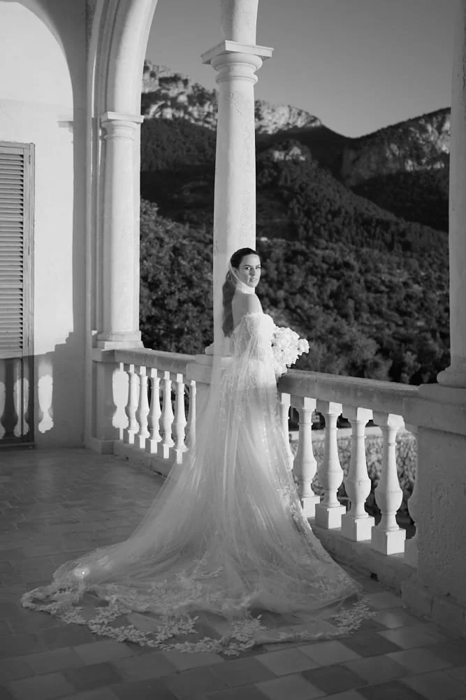 A woman in a wedding dress holding a bouquet of flowers standing on a balcony with classical columns, overlooking mountains and trees in the background.