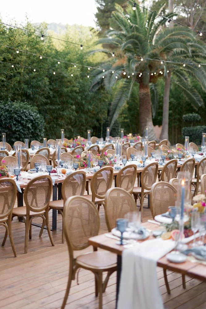 Outdoor event setup with long tables, wooden chairs, string lights, and tropical greenery in the background.