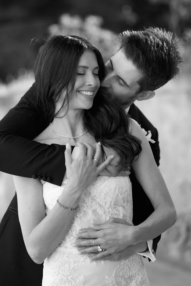 A black and white photo of a couple embracing, with the man kissing the woman's cheek. The woman is smiling with her eyes closed, wearing a wedding dress. The man is wearing a suit, and they are both showing rings on their fingers.