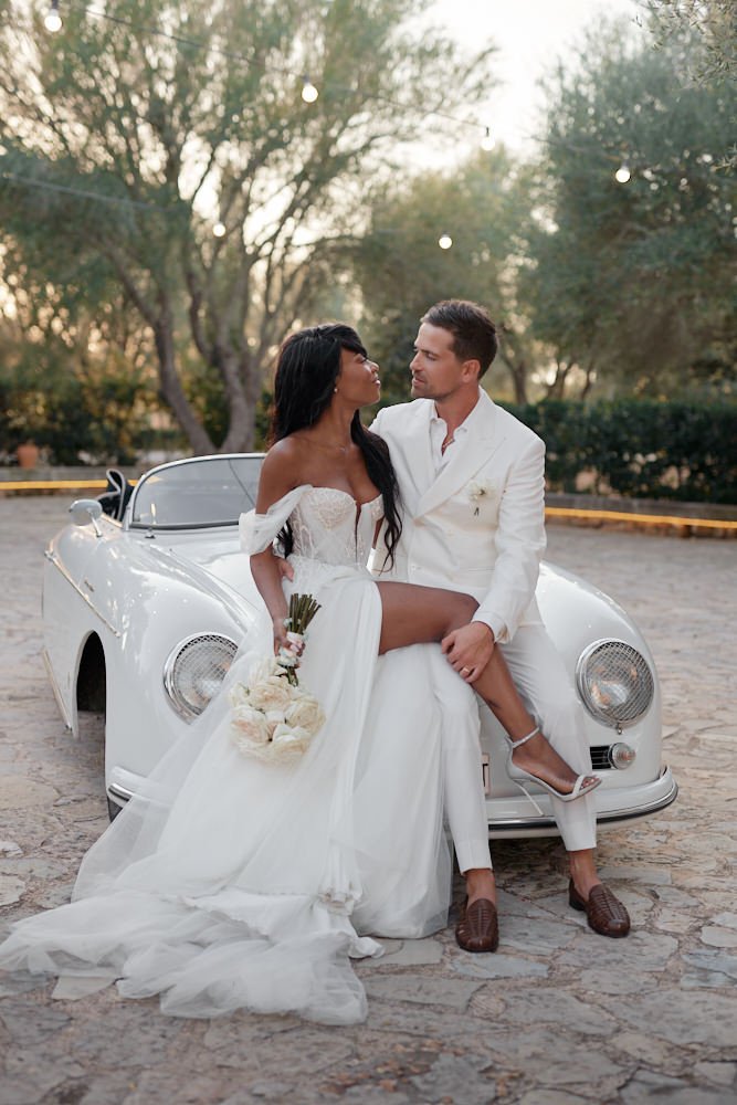 A couple dressed in wedding attire sitting on the front of a vintage white sports car outdoors with trees in the background.