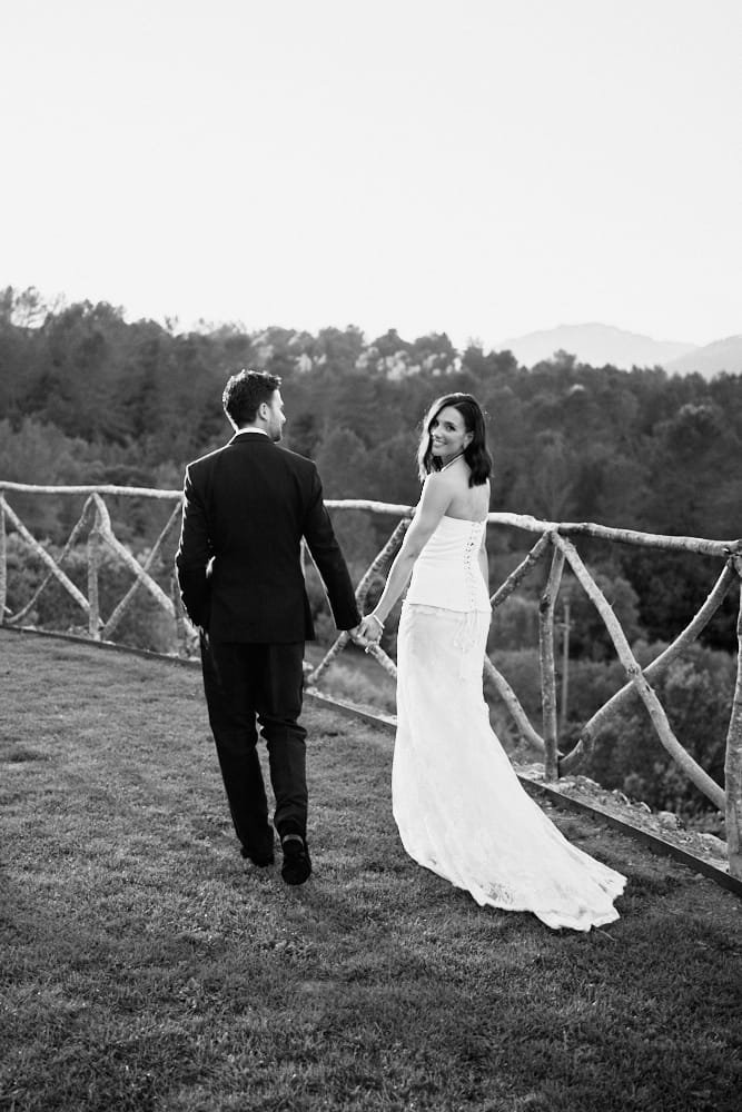 A woman in a white wedding dress and a man in a black suit walking hand in hand outdoors, with a scenic landscape and trees in the background.