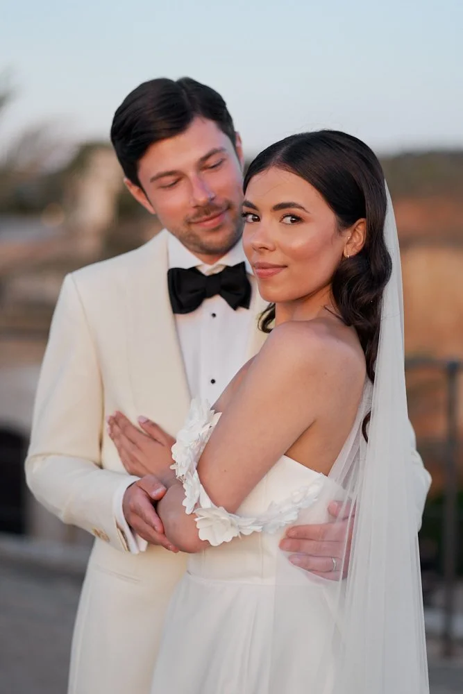 A bride and groom in wedding attire embrace outdoors, with the groom in a white tuxedo with a black bow tie and the bride in a white wedding dress with off-the-shoulder details, posing at sunset.
