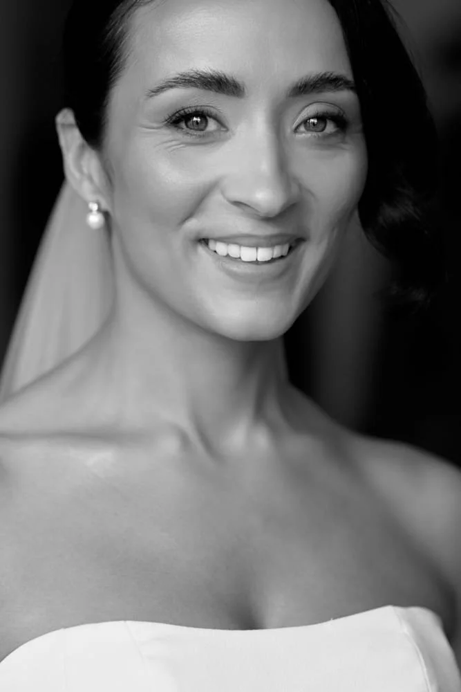 Close-up portrait of a smiling woman in a wedding dress with pearl earrings, black and white photography.