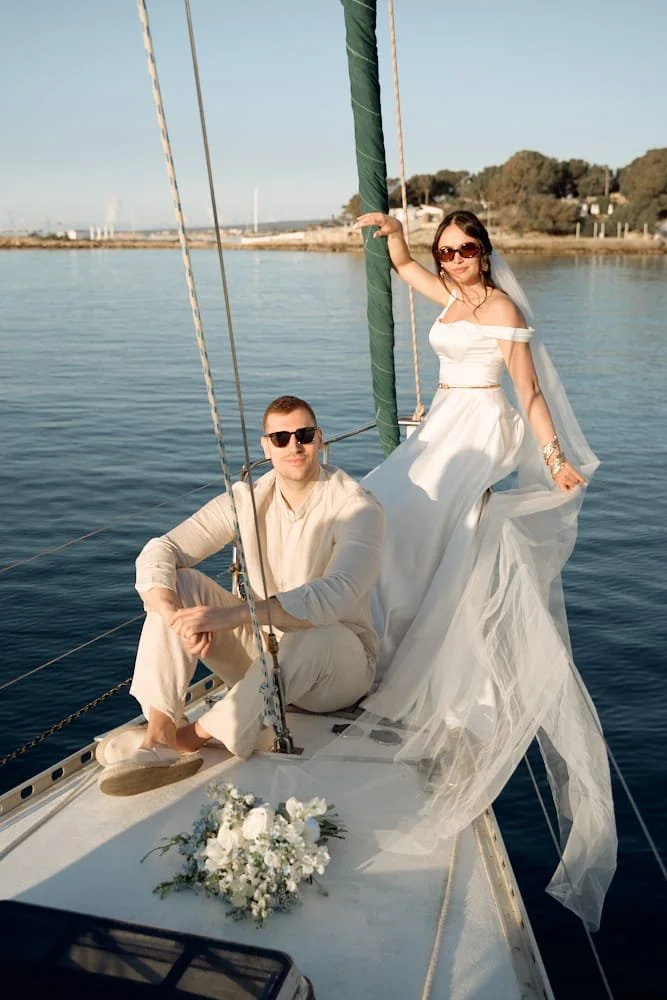 A woman in a white wedding dress and a man in light-colored clothing sitting on a sailboat, with the woman standing and holding onto the sail mast, both wearing sunglasses, with a bouquet of flowers on the boat and water in the background.