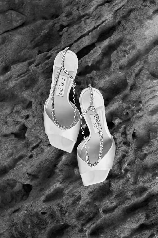 White high-heeled sandals with rhinestone straps lying on a rocky surface.