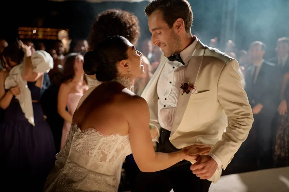 A bride and groom dance closely at their wedding reception, surrounded by guests in a dimly lit venue.