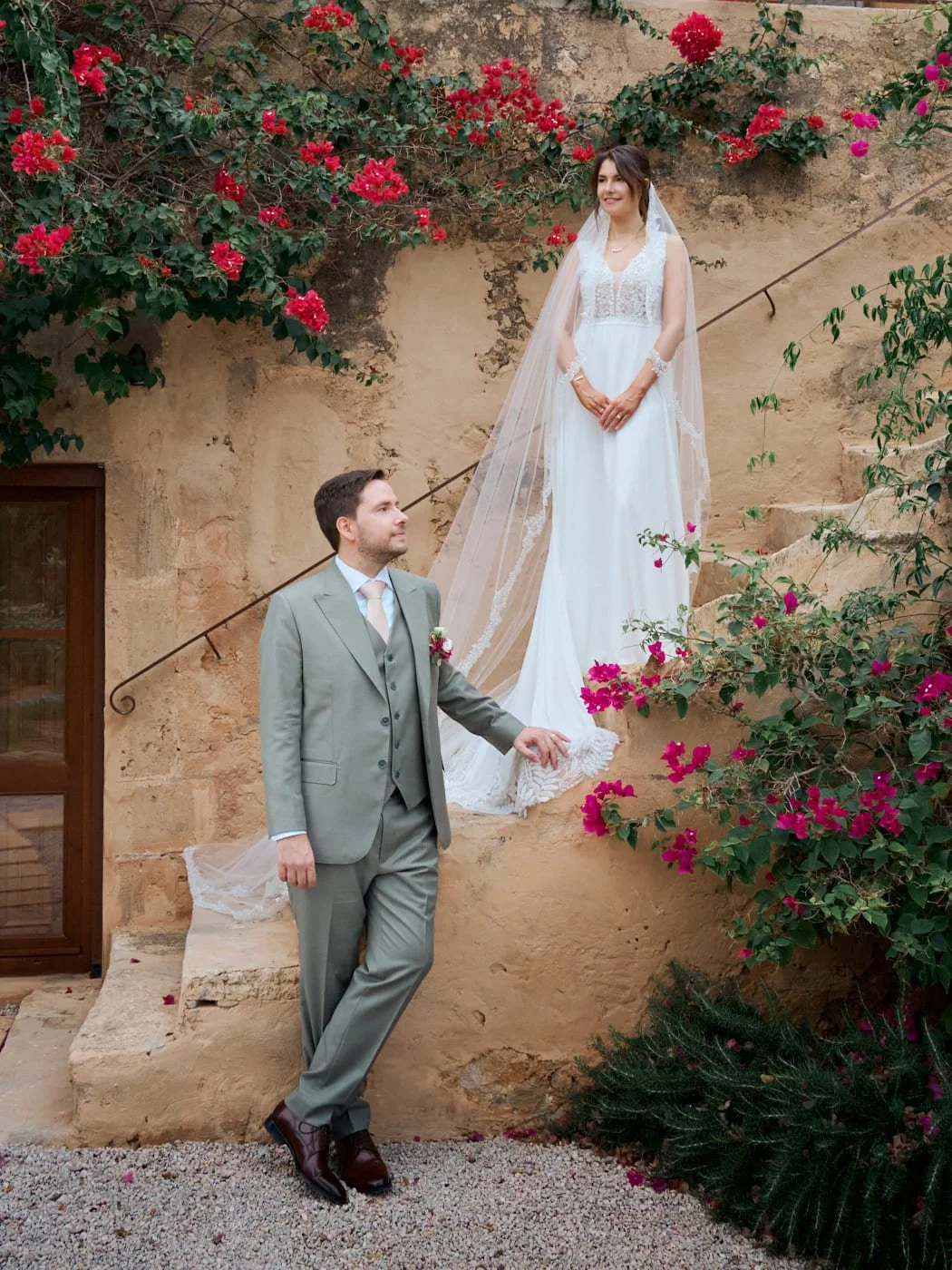 A bride in a white wedding dress and veil stands on stone stairs surrounded by pink and red flowers, looking happy, while a groom in a gray suit and tie stands below, looking up at her in a garden setting. Wedding in Mallorca. The Lumery.