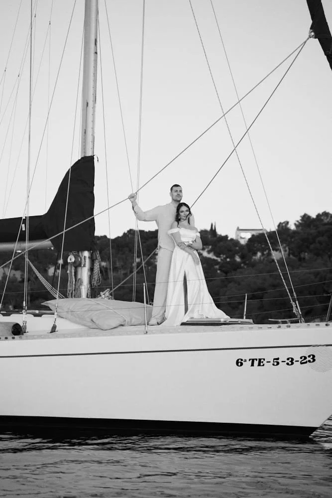 A couple stands on a sailboat, with the man behind the woman, holding her. The woman holds a small dog. The boat is on calm water with a shoreline and trees in the background. The image is in black and white.