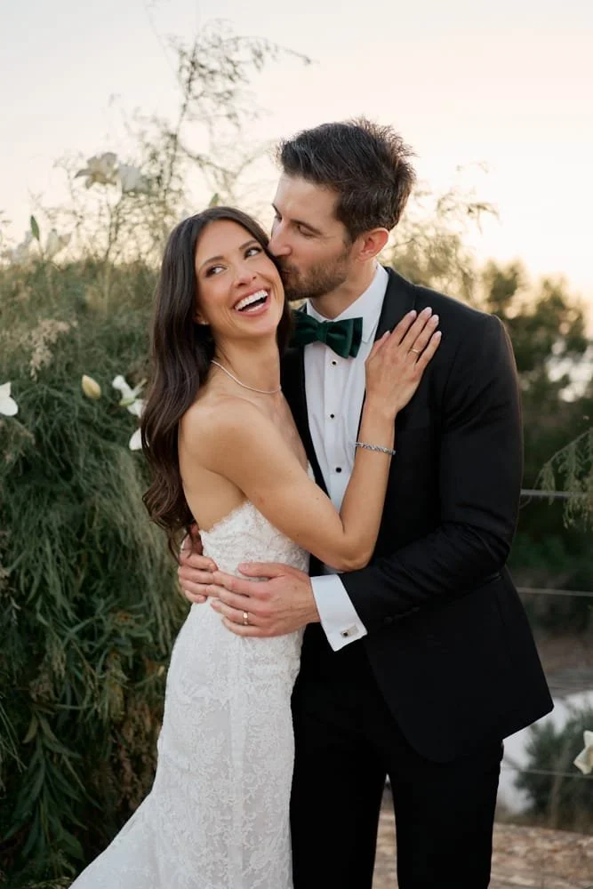 A newlywed couple, dressed in wedding attire, embracing outdoors at sunset, surrounded by greenery.
