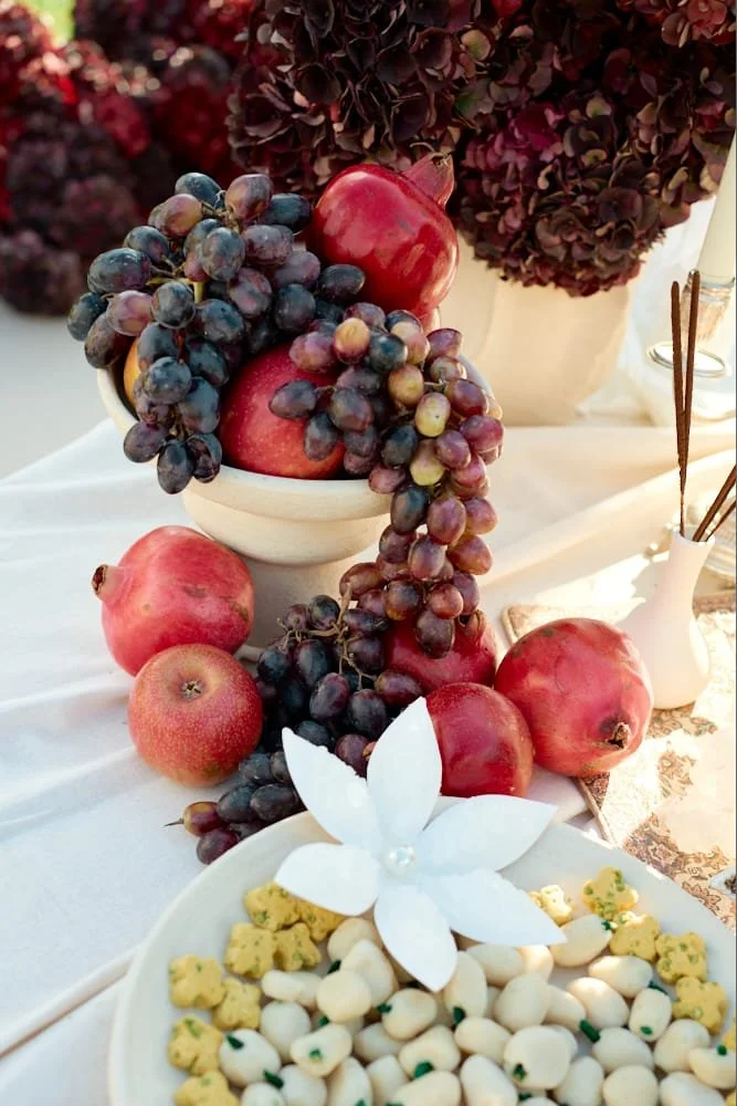 A table setting with red apples, green grapes, and white and yellow confections arranged around a white tablecloth and a large floral centerpiece with dark purple hydrangeas.
