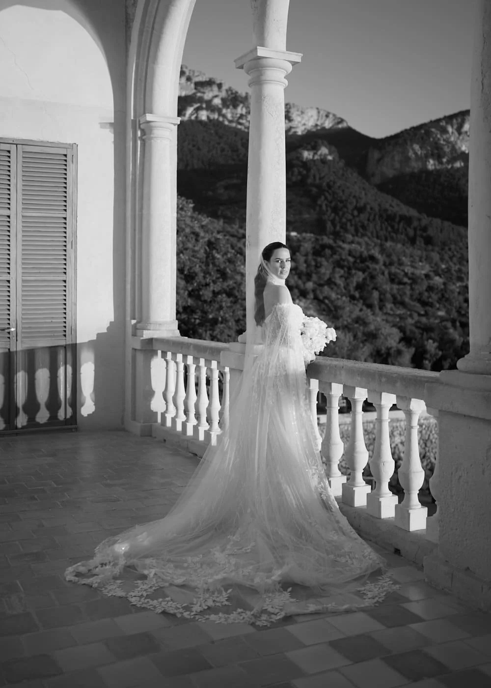 A bride in a wedding dress holding a bouquet on a balcony with ornate columns, overlooking mountains in the background. Son Marroig.