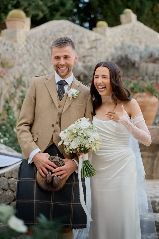 A newlywed couple at their wedding, smiling and happy. The groom is in traditional Scottish attire, wearing a kilt and holding a sporran. The bride is in a white wedding dress, holding a bouquet of white flowers, and appears joyful.