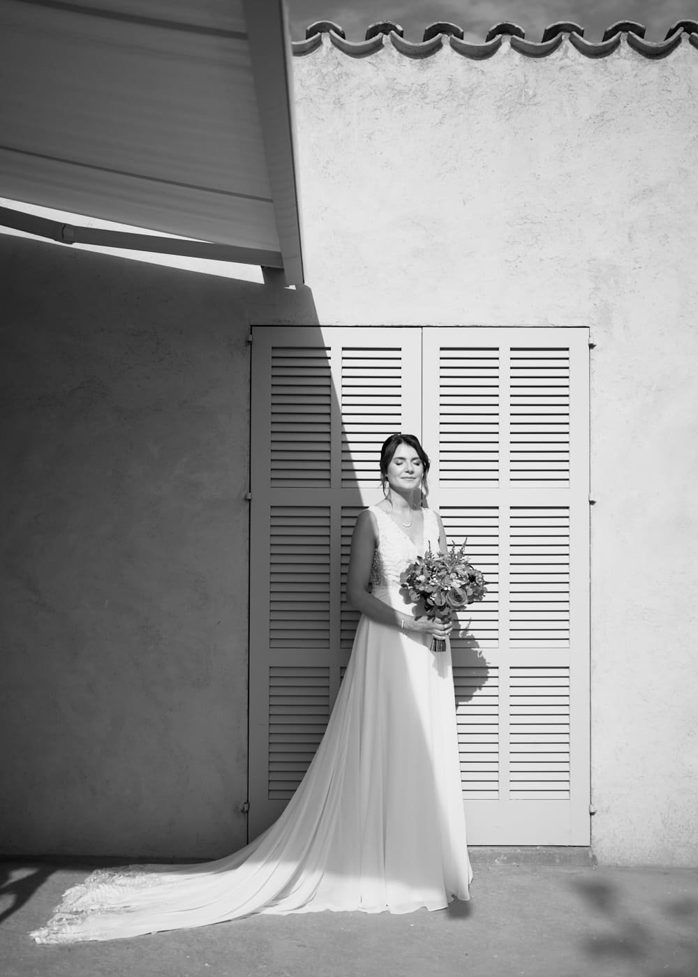 A bride in a white wedding gown standing outdoors, holding a bouquet of flowers, with her eyes closed and a slight smile, sunlight casting shadows on her face and background. Wedding in Mallorca. The Lumery.