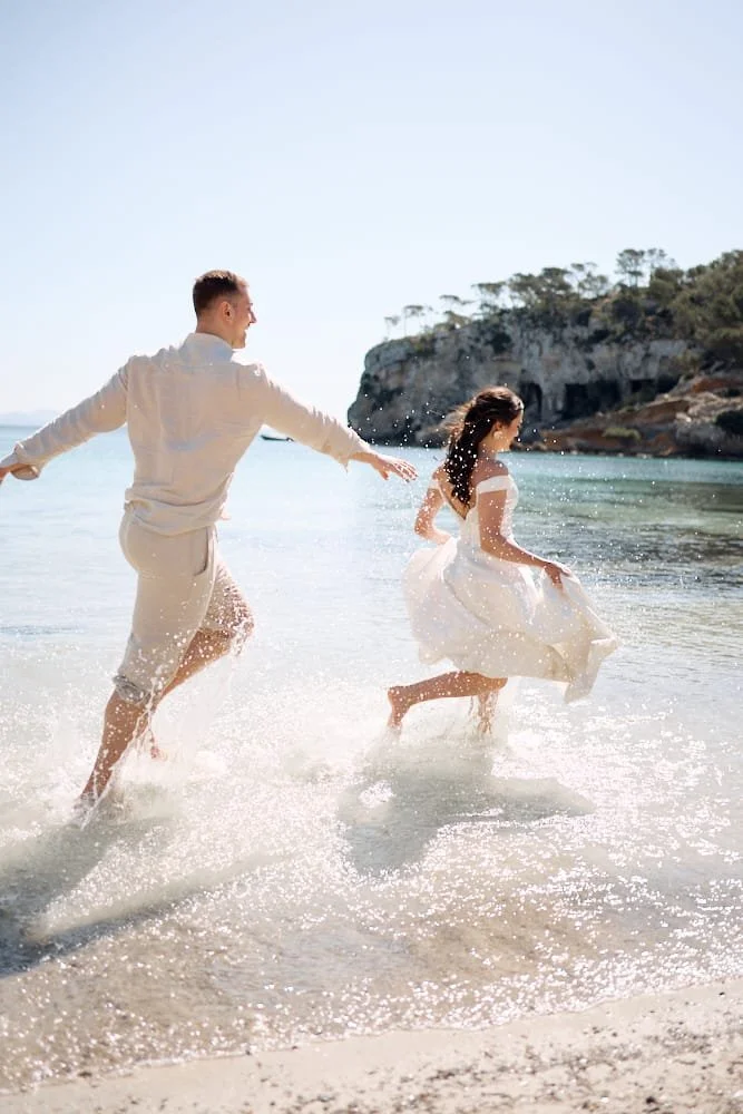 A man and woman running and playing in shallow water at the beach, with cliffs and trees in the background.