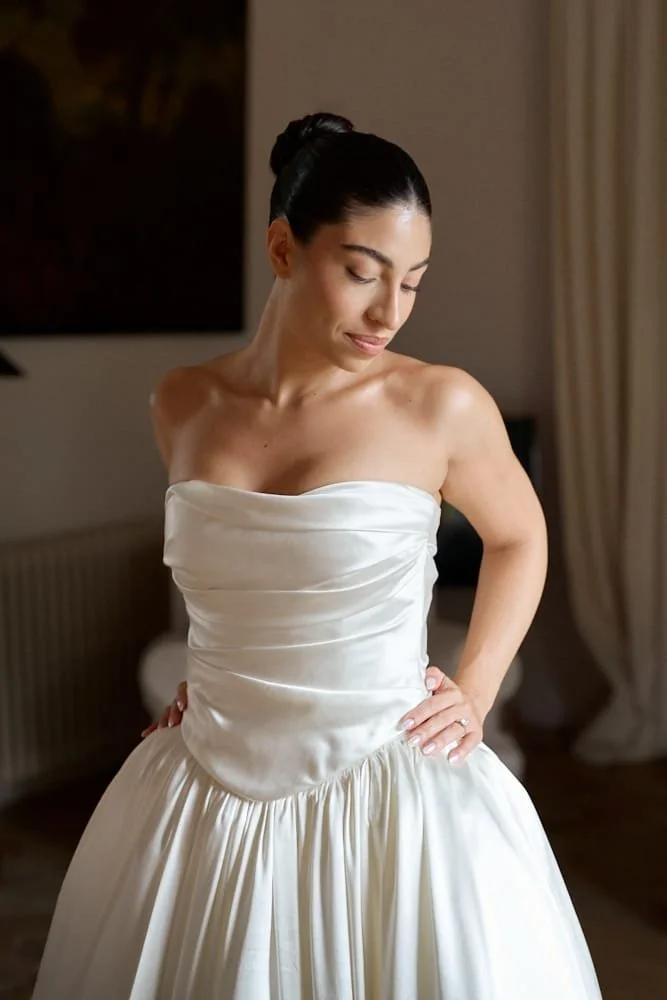 A woman in a strapless white gown with a ruched bodice and full skirt, standing indoors with her eyes closed and hands on her hips.
