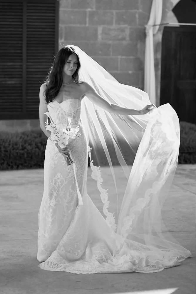 A bride in a strapless lace wedding gown holding a bouquet of flowers stands outdoors, adjusting her long veil with one hand, near a brick building and shrubbery.