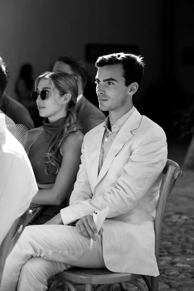 A man in a light-colored suit sitting at a formal event with other attendees around him, including a woman in sunglasses.
