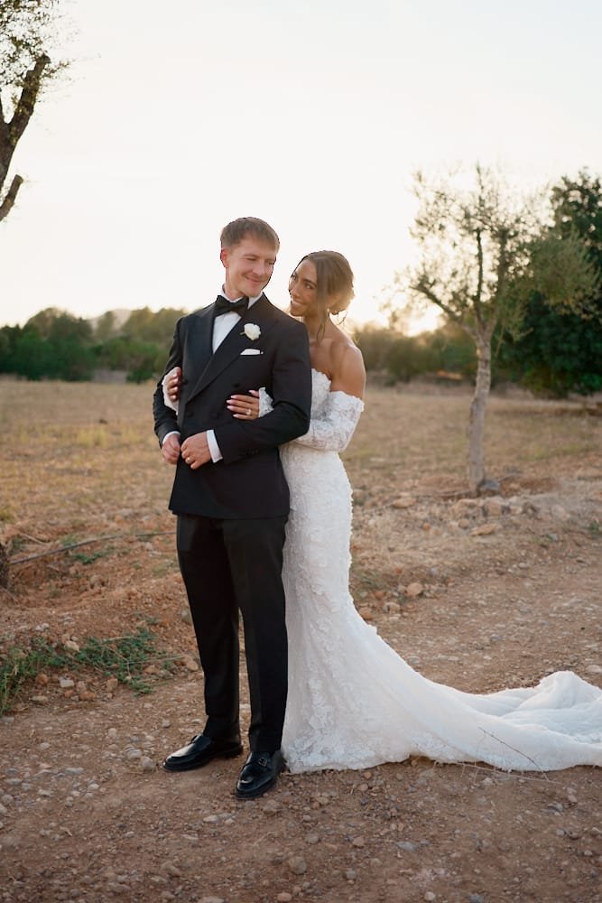 A bride and groom stand outdoors on a dirt path, smiling and looking at each other during sunset. The groom wears a black tuxedo with a white shirt and bow tie, and the bride wears an off-the-shoulder white wedding gown with a train.
