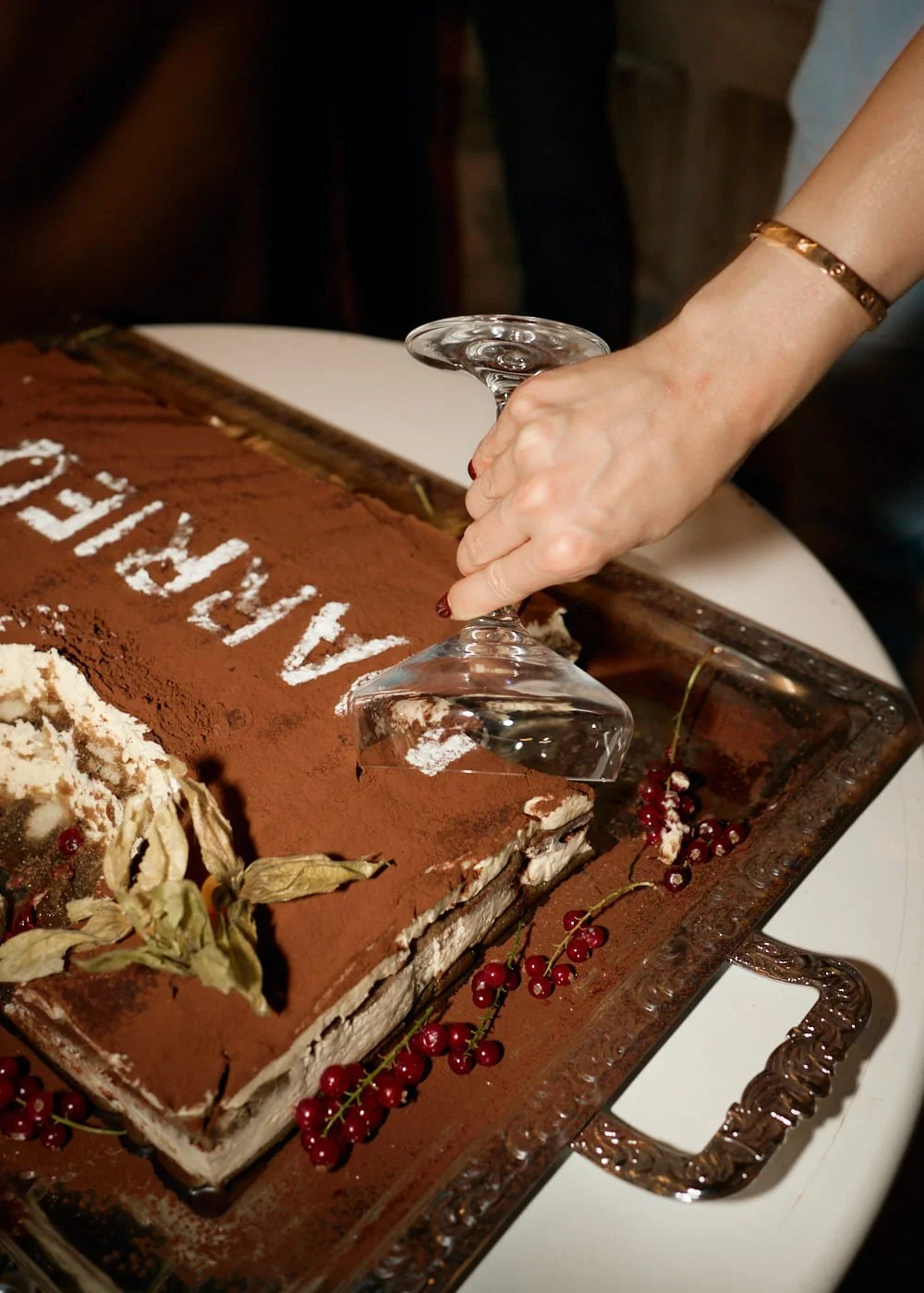 Person holding a wine glass upside down above a large rectangular layered dessert cake with cocoa powder, red currants, and edible leaves, on a decorative platter. Wedding in Mallorca. The Lumery.