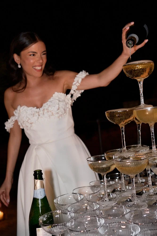 A woman in a white dress with floral details on the shoulder is pouring champagne into a stacked pyramid of champagne glasses.