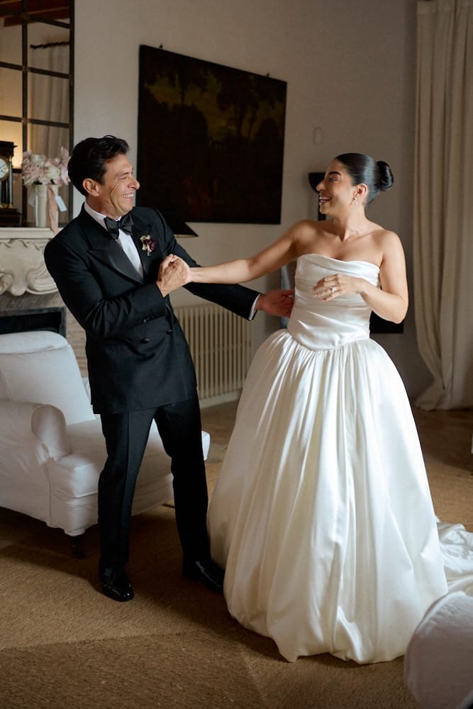 A bride in a strapless white wedding gown and an older man in a tuxedo holding hands and smiling in a warmly lit living room.