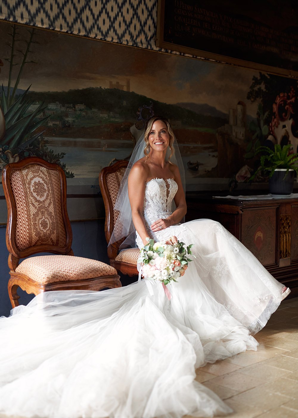 Bride in white lace wedding gown sitting on chair, holding bouquet, with veil and long train, in ornately decorated room with a mural.
