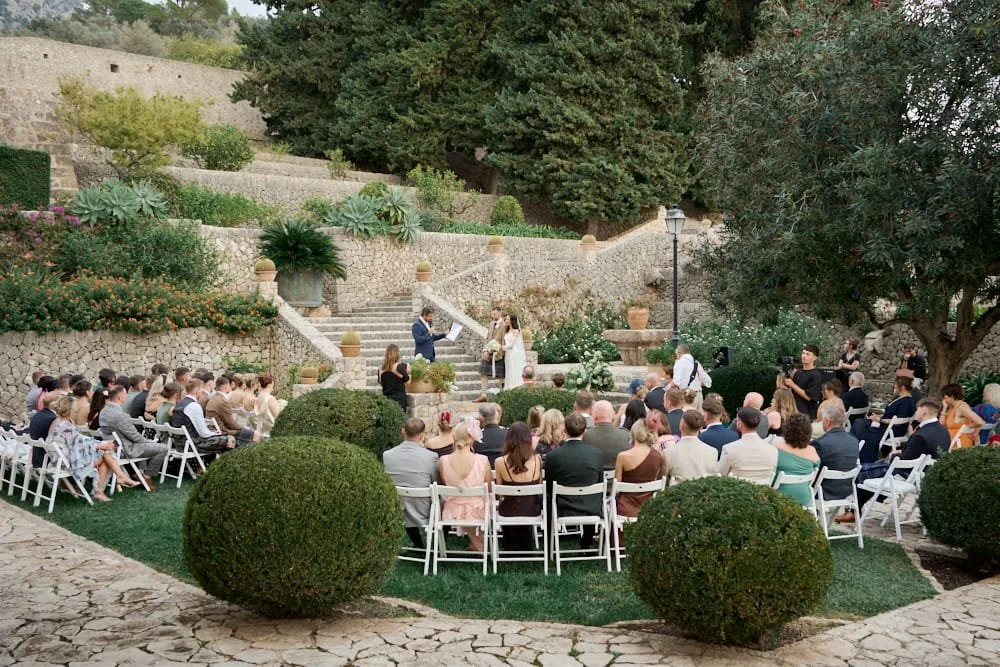 Outdoor wedding ceremony with guests seated in white chairs, couple exchanging vows on stone stairs, surrounded by lush greenery and landscaping.