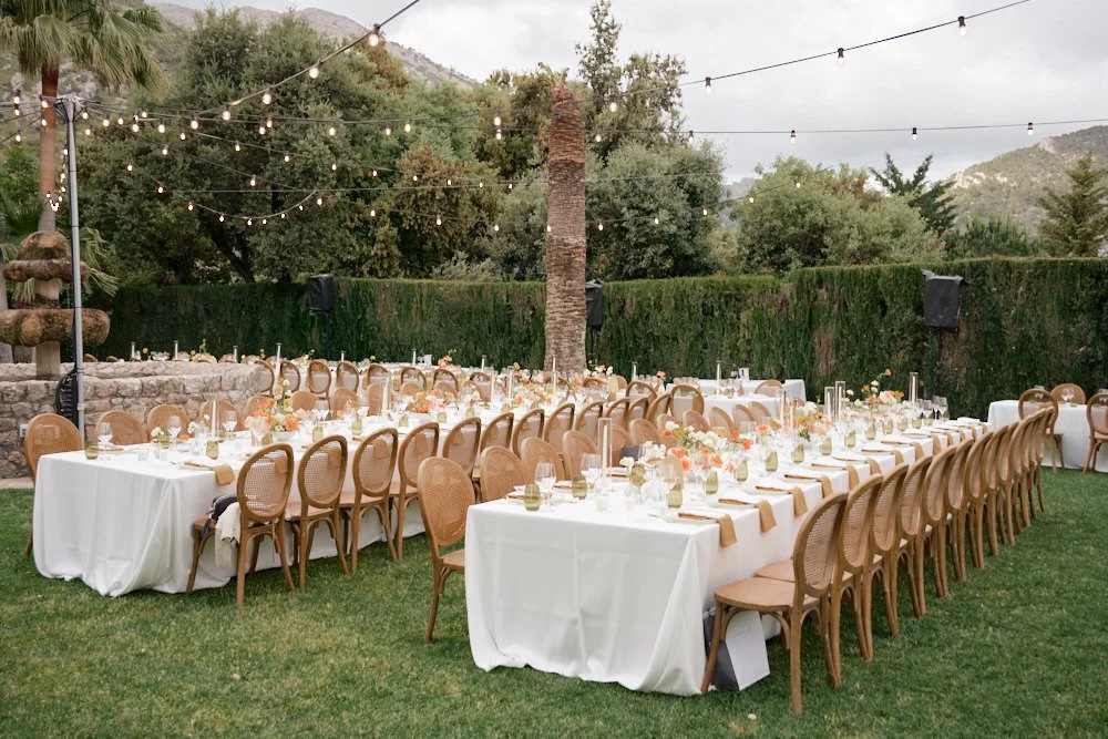 Outdoor wedding reception setup with long rectangular tables covered with white tablecloths, decorated with flowers, candles, and place settings, surrounded by wooden chairs, under string lights with a cloudy sky over mountains and trees in the backg