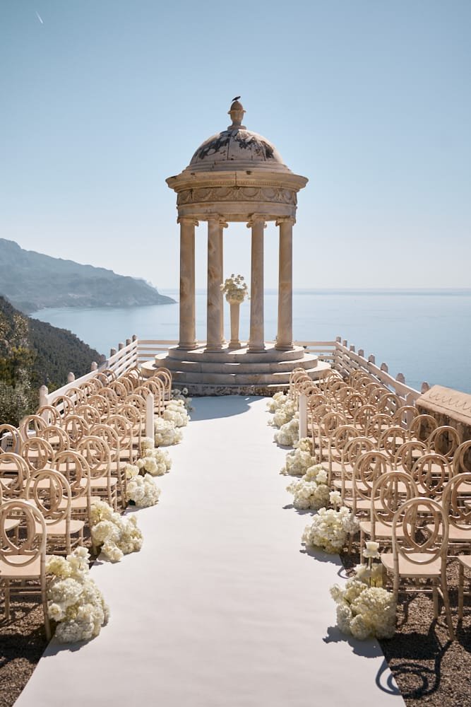 Outdoor wedding ceremony setup with a white aisle, surrounded by white floral arrangements, leading to a classical gazebo overlooking the ocean with mountains in the background.