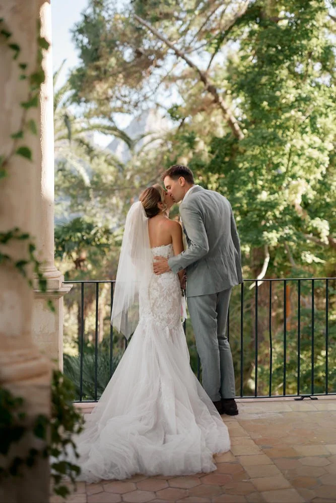 A bride and groom share a kiss on a balcony surrounded by trees, with the bride in a white wedding gown and veil, and the groom in a gray suit.