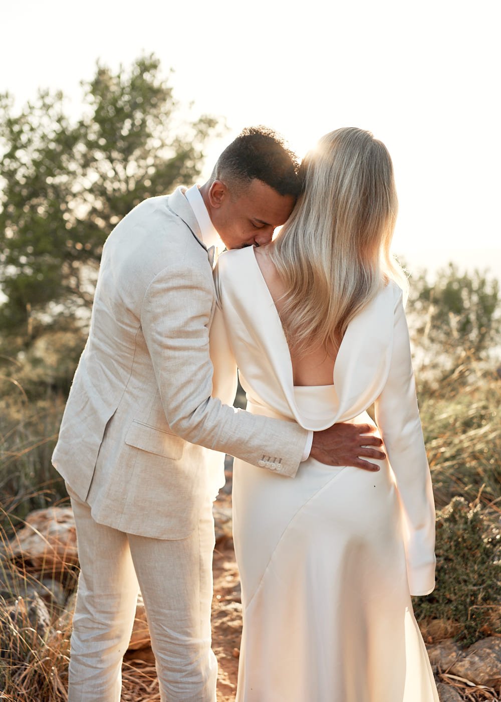 A couple embracing outdoors in formal attire, with the man in a light suit kissing the woman's shoulder as she wears an open-back white dress. The background features trees and soft sunlight.