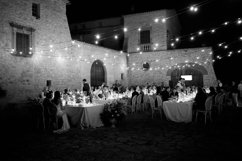 Outdoor nighttime wedding reception with guests seated at long tables under string lights, held in a stone courtyard surrounded by historic stone buildings.