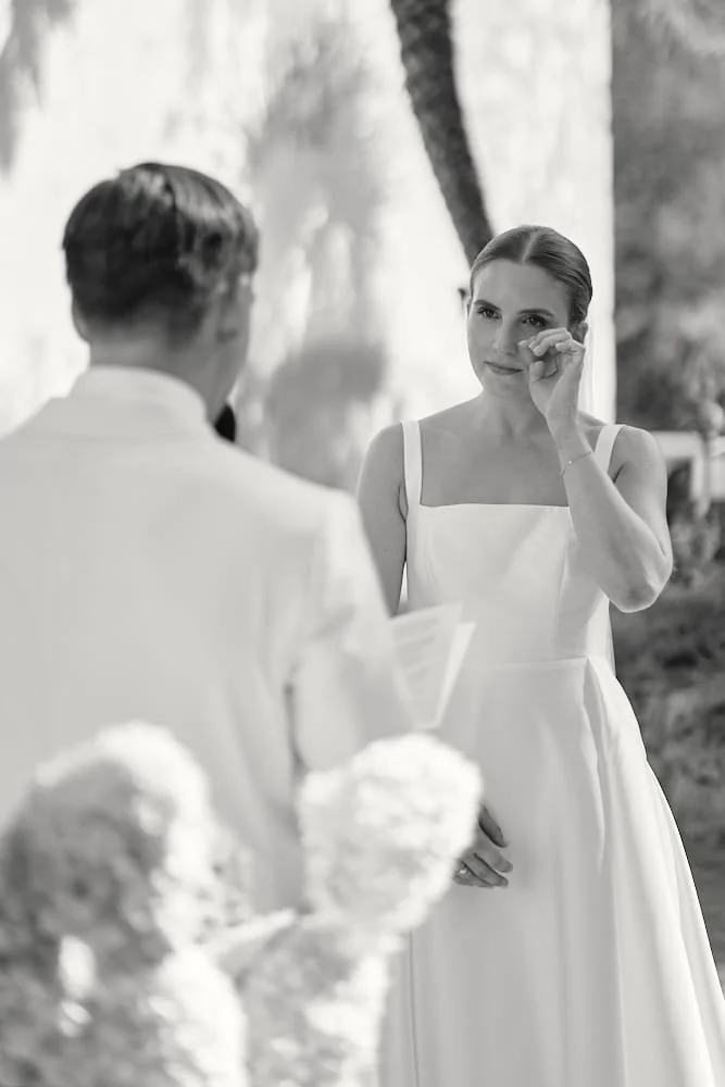 A woman in a white dress wiping away tears while looking at a man in a white shirt during a wedding ceremony outdoors.