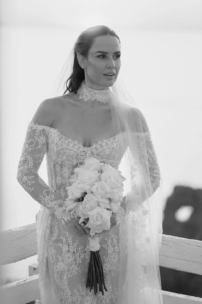 Black and white photo of a bride in a lace wedding dress holding a bouquet of roses, standing outdoors with a railing and natural scenery in the background.