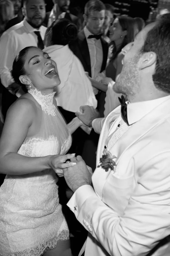 A wedding reception scene in black and white. A bride and an older man, likely her father, are dancing and smiling. The bride is wearing a lace wedding dress, and the man is in a tuxedo with a boutonniere. People in the background are dressed formall