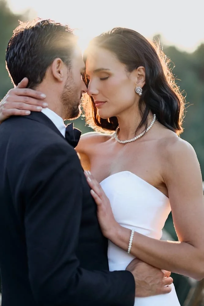 A bride and groom with foreheads touching outdoors at sunset, embracing lovingly, dressed in wedding attire.