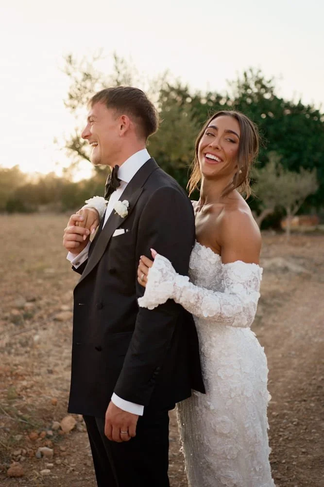 A bride and groom standing outdoors, both smiling and laughing. The bride is wearing a white lace off-the-shoulder wedding dress, and the groom is in a black tuxedo with a bow tie. The couple appears happy and affectionate.