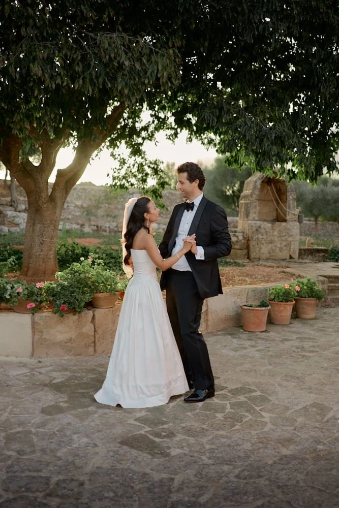A bride and groom dancing outdoors under a large tree, with sunlight filtering through the leaves, surrounded by potted plants and rustic stone structures.