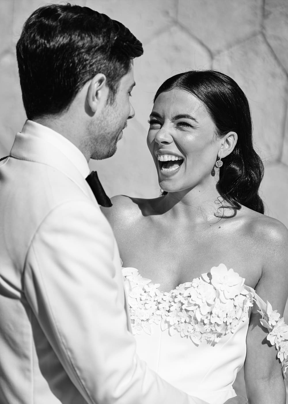 A black and white photo of a couple smiling and embracing, the woman wearing an embellished wedding dress and the man in a suit.