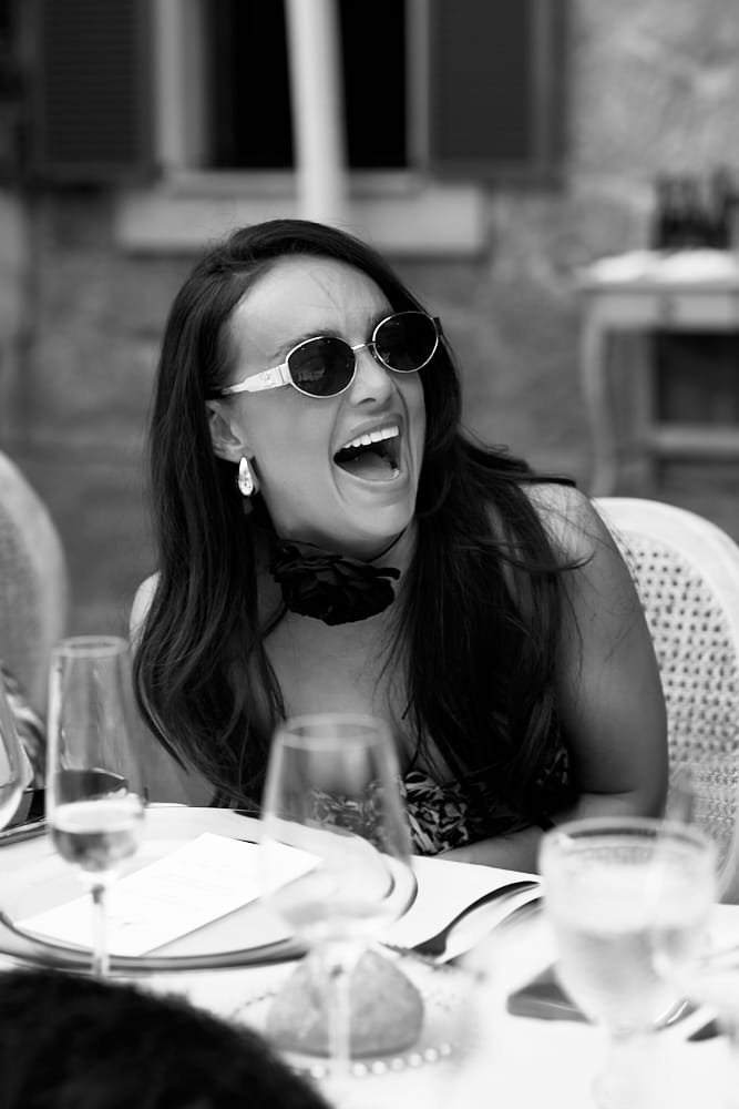 Woman with long dark hair, wearing sunglasses, earrings, and a floral dress, laughing at a dinner table.