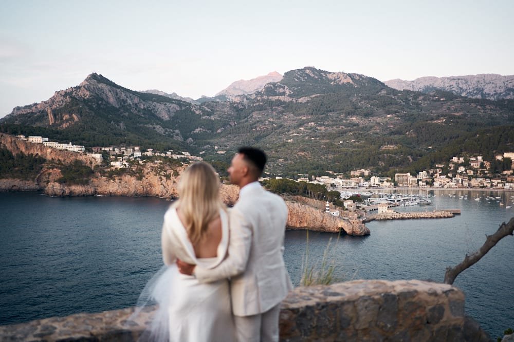 A couple in wedding attire standing on a scenic overlook by the water, with mountains and a coastal town in the background.