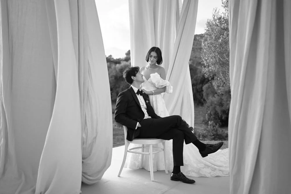 Black and white photo of a woman in a wedding dress holding a baby, standing next to a man in a tuxedo sitting on a chair, with curtains and an outdoor landscape in the background.