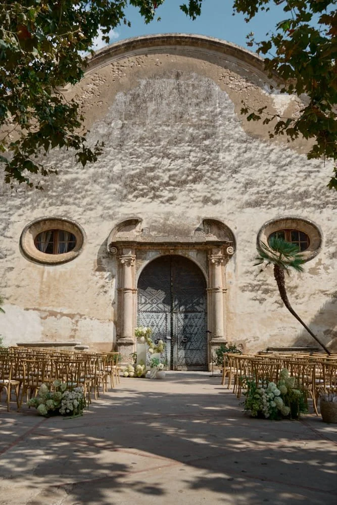 Outdoor wedding ceremony setup with wooden chairs and floral arrangements in front of an old stone building with a large arched door and two round windows, shaded by trees.