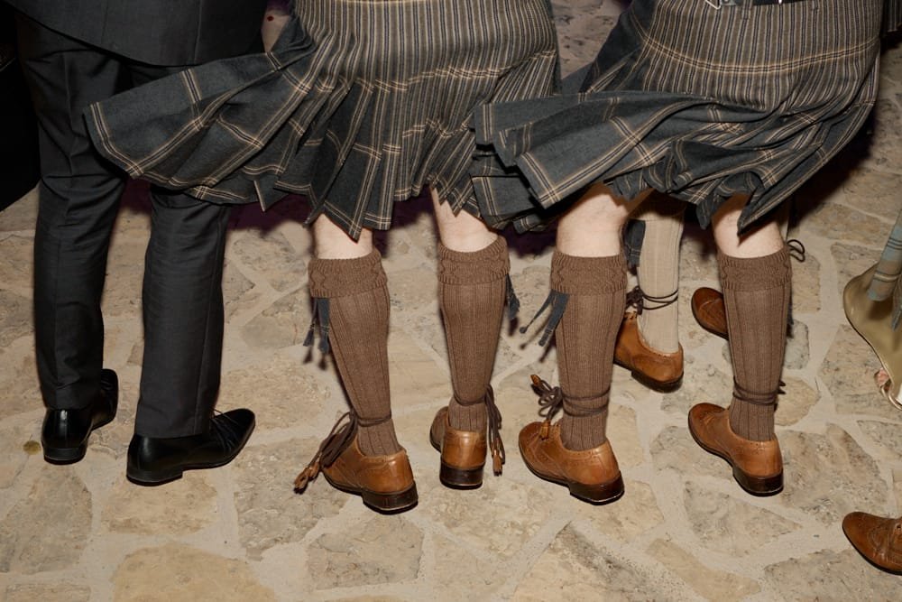 Three people wearing kilts with knee-high socks and brown shoes, seen from the back, sitting on stools.