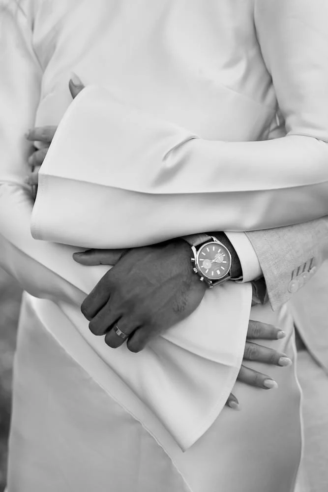Close-up of a person's arm and hand crossing in front of another person's arm, with a large wristwatch on the wrist, in black and white.