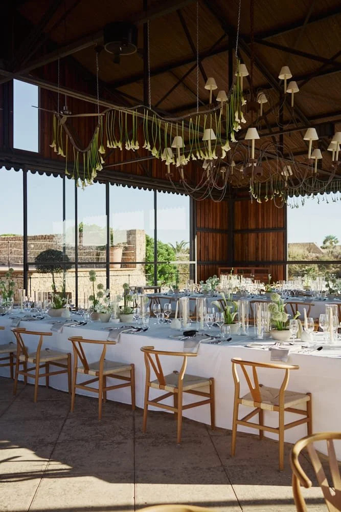 Elegant outdoor dining setup with a long table covered in a white tablecloth, decorated with white flower arrangements and candles, set with glassware, plates, and silverware. Wooden chairs surround the table, inside a wooden pavilion with large glas