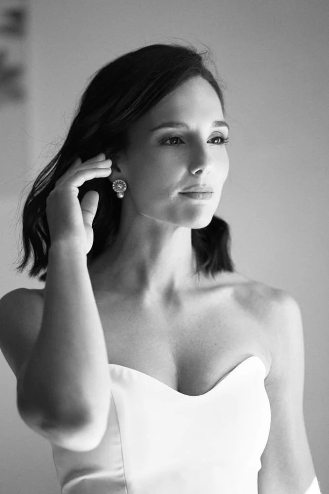 Black and white portrait of a woman with shoulder-length dark hair, wearing pearl earrings and a strapless dress, touching her ear with her left hand, gazing slightly to the side.